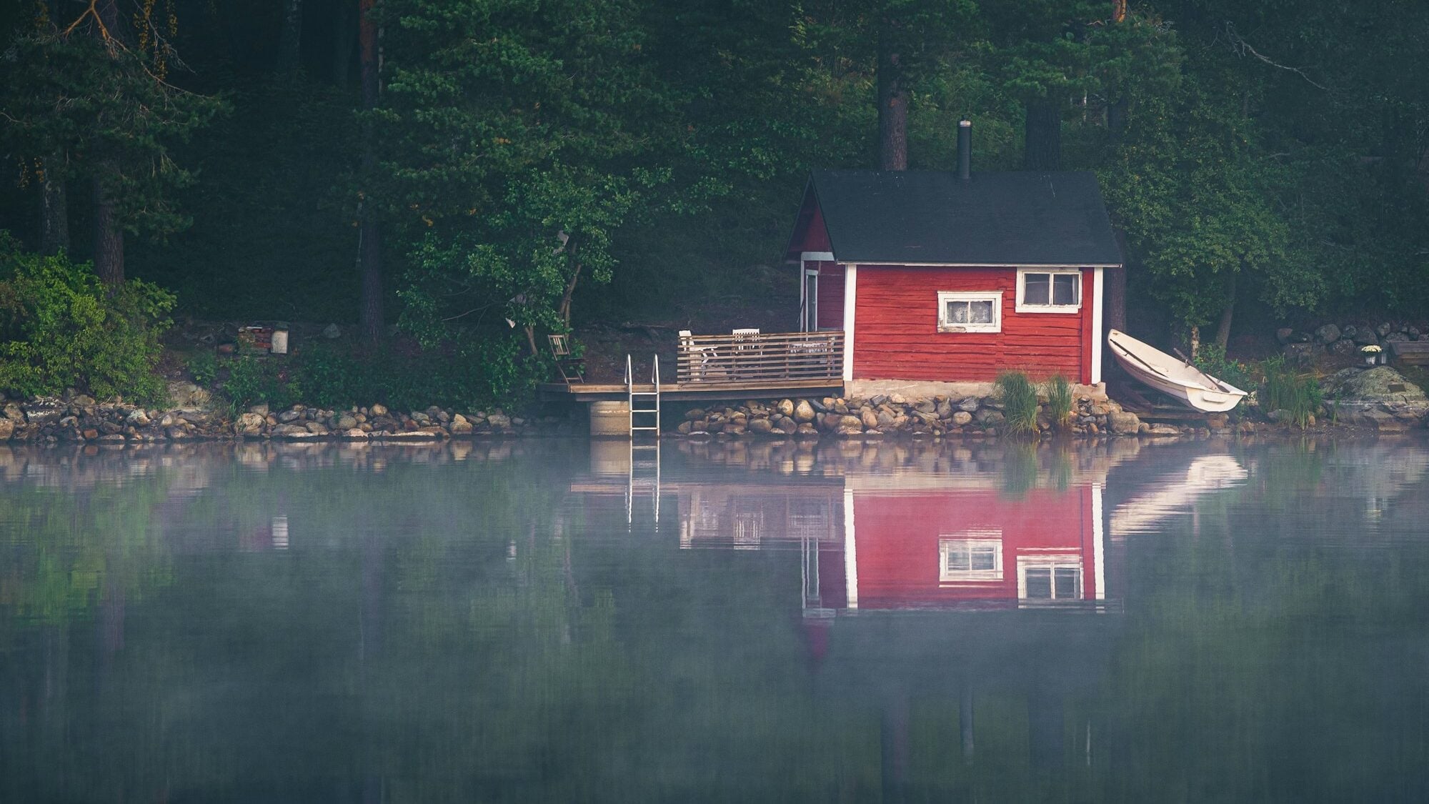Punainen sauna- tai mökkirakennus järven rannalla metsän reunassa aamusumussa