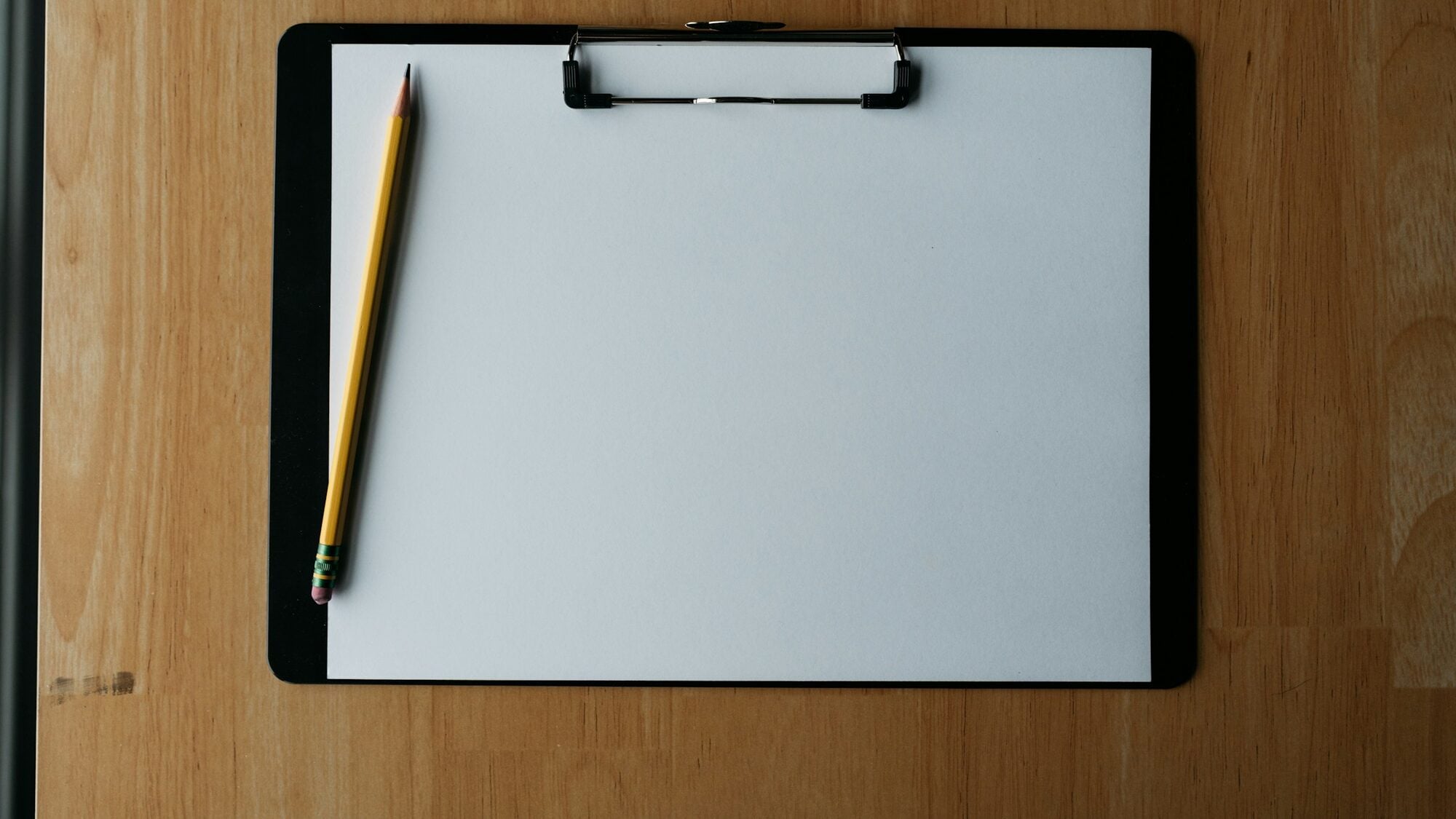 A blank sheet of paper on a clipboard with a pencil on a wooden desk